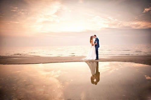 Far shot of bride and groom standing and facing one another on reflective sand in front of the ocean