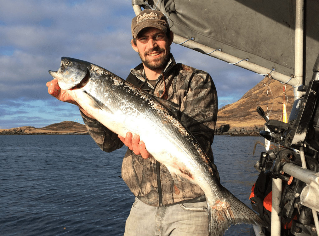 close-up of a young bearded man wearing a Cabela's baseball cap and thin camo jacket smiling while holding a large fish on a boat