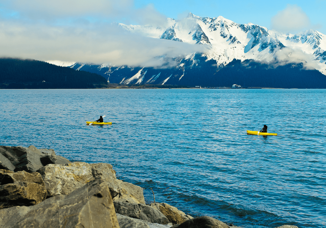 two kayakers paddling on a clear blue lake in alaska in front of a snow-covered mountain partially obscured by clouds
