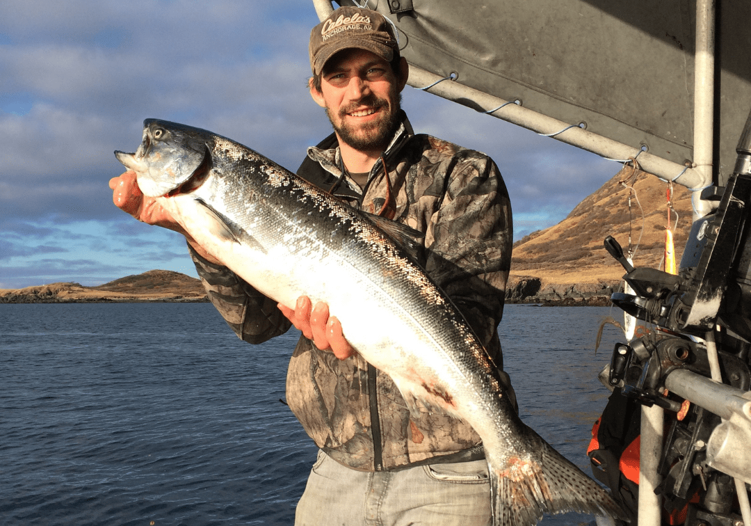 close-up of a young bearded man wearing a Cabela's baseball cap and thin camo jacket smiling while holding a large fish on a boat