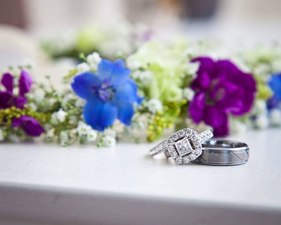 Silver his and her wedding rings set together in front of a wildflower wreath with violet flowers and baby’s breath